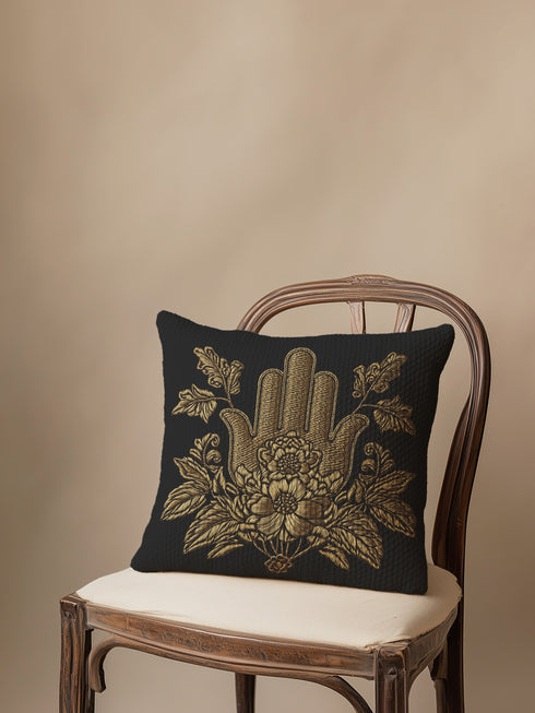 A black woven throw pillow featuring an embroidered gold Hamsa hand surrounded by intricate floral and botanical designs.